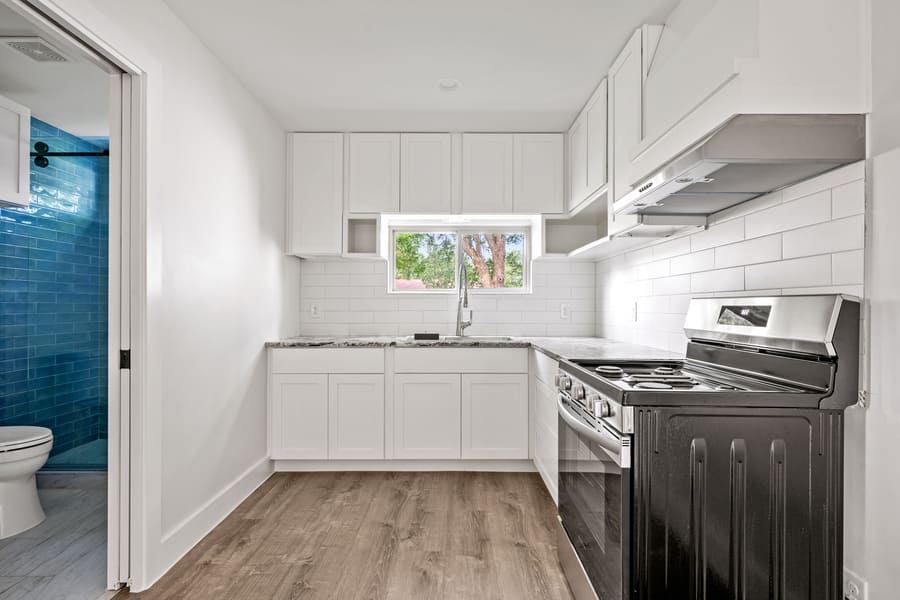 Small modern kitchen with white cabinets, granite countertops, stainless steel stove, and a window above the sink; adjacent bathroom with blue subway tiles and a toilet visible.