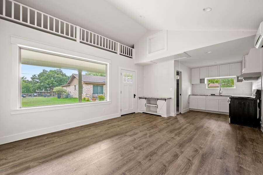 Bright, empty living space with large window, wood floor, white walls, and a small open kitchen with white cabinets and black appliances.