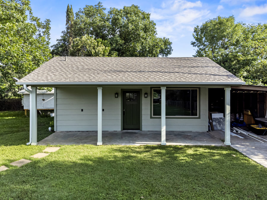 Small single-story house with gray siding, green door, large window, and a covered front porch with four white columns.