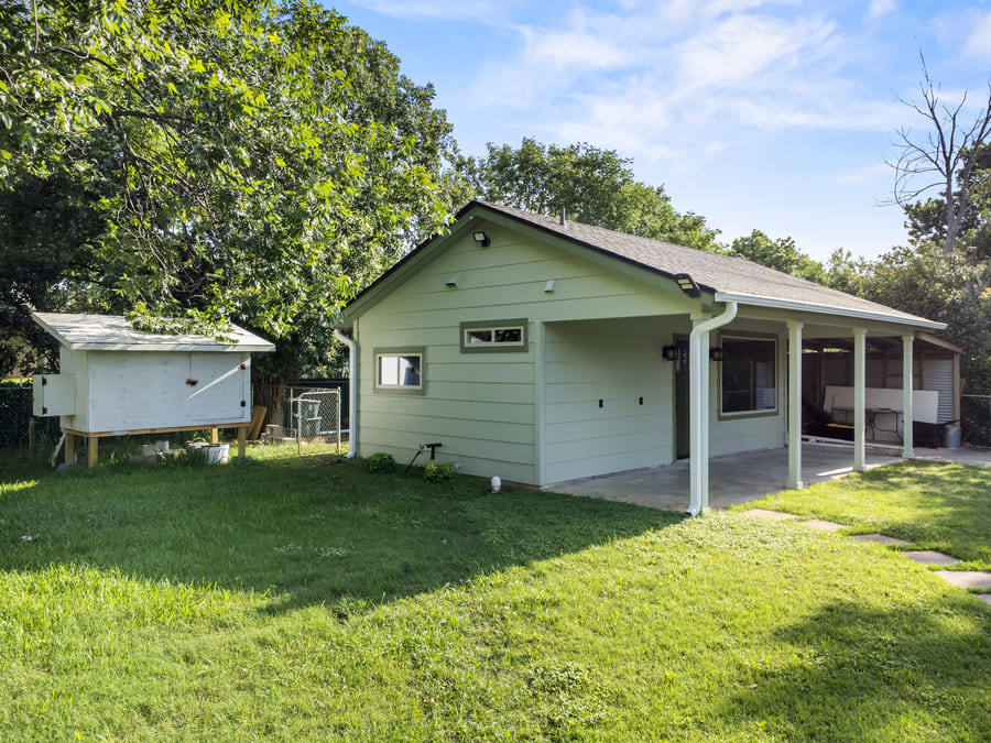 Small pale green house with a covered porch in a green yard with trees and a wooden enclosure to the left.