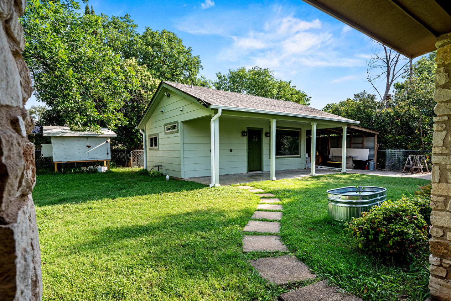 Small light green house with a covered porch and green door, surrounded by a grassy yard with stepping stones, trees, and a metal water trough.