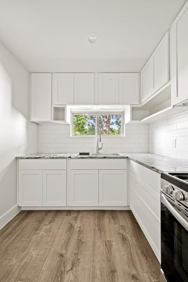 Bright modern kitchen with white cabinets, marble countertops, stainless steel sink, and a window overlooking green trees.