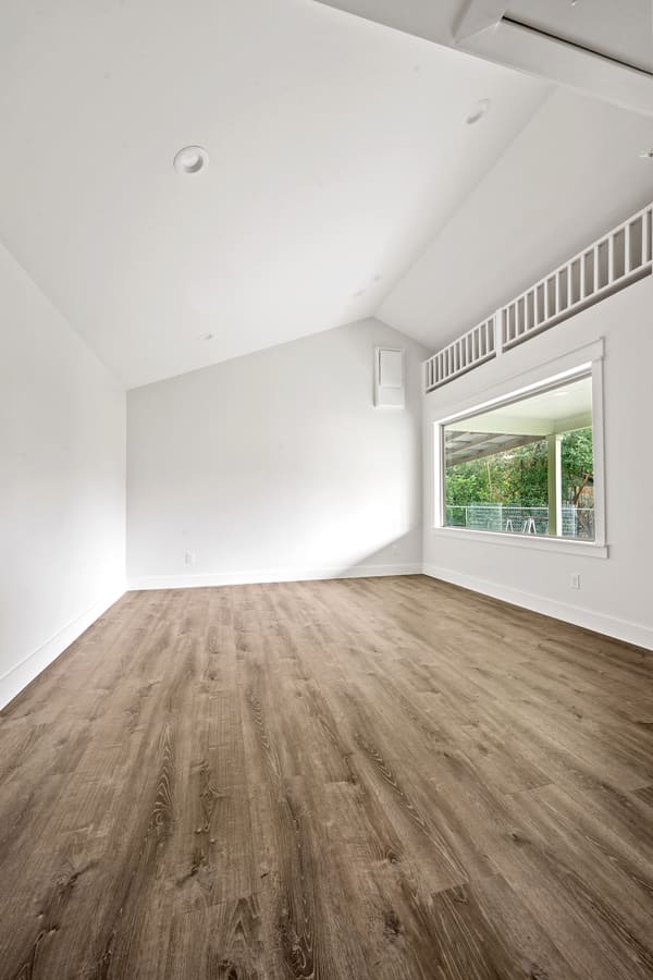 Empty room with vaulted white ceiling, wood-look flooring, and a large window showing a green outdoor area.
